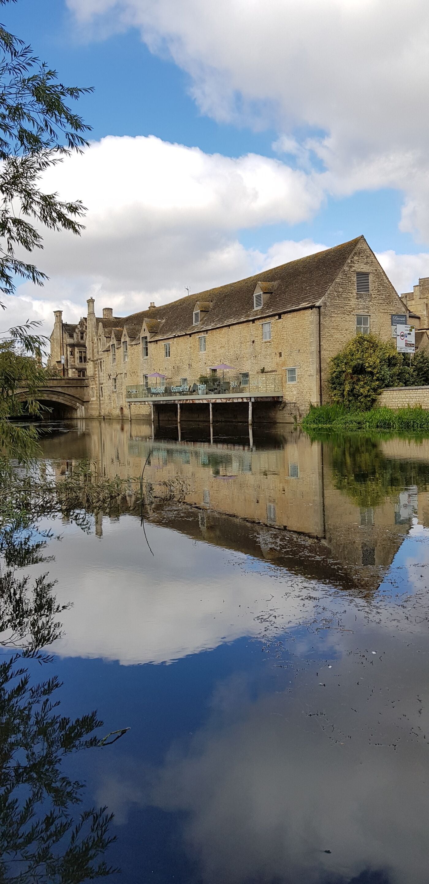 Ein historisches Steinhaus mit einer Brücke über einem ruhigen Fluss, der Gebäude und Himmel spiegelt.