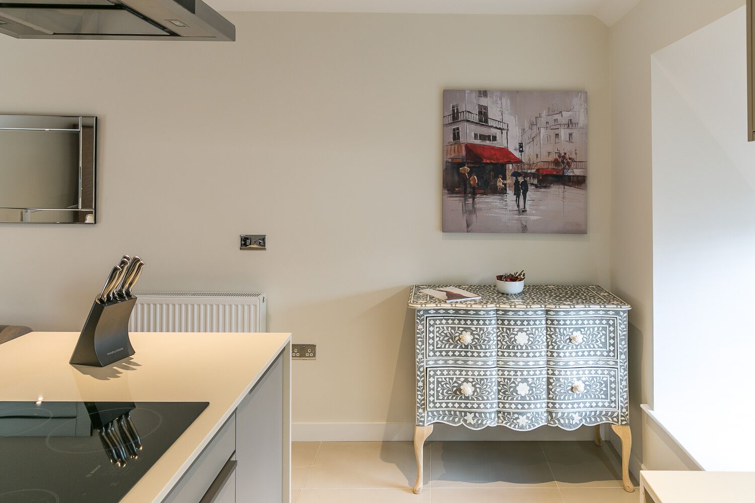 A modern kitchen with a white countertop, a black knife holder, a patterned cabinet, and a cityscape painting on the wall.