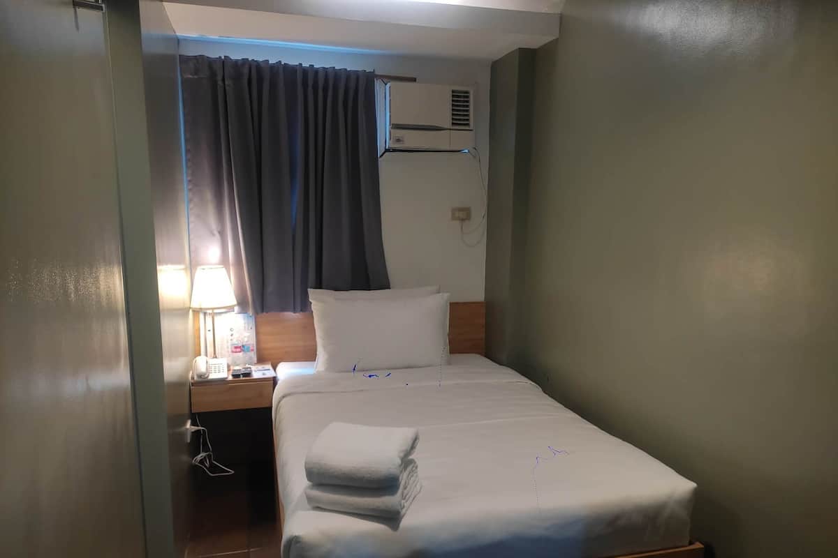 Un petit lit bien fait, avec du linge de lit blanc et une serviette pliée, une lampe de chevet, une table de chevet avec un téléphone et une bouteille, et une fenêtre avec des rideaux.