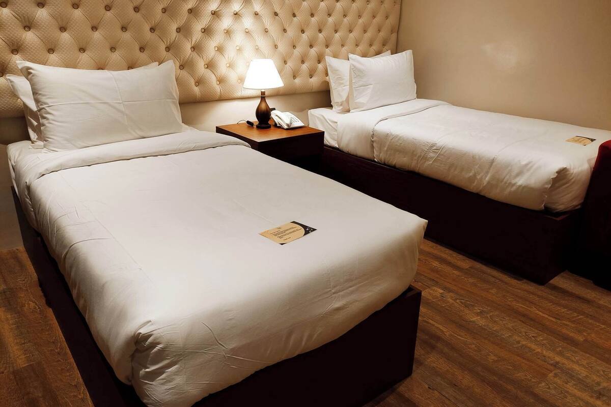 Une chambre d’hôtel avec deux lits simples, une table de chevet en bois avec une lampe et une tête de lit capitonnée.