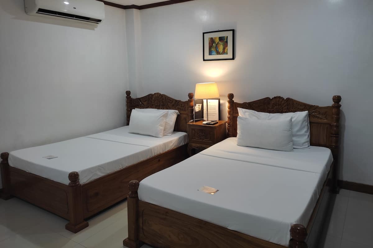 Une chambre d’hôtel avec deux lits en bois, une table de chevet avec une lampe, un climatiseur et un tableau encadré au mur.