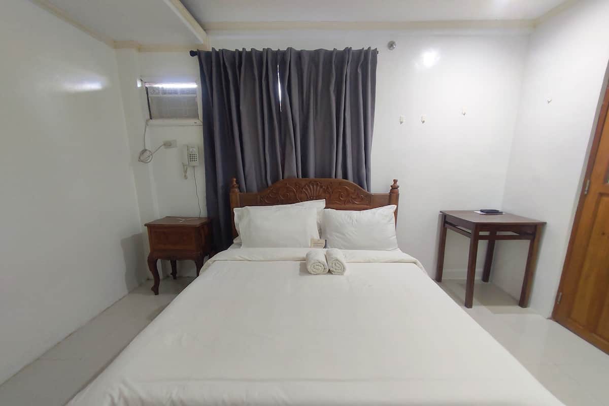 Un lit bien fait, avec du linge de lit blanc et deux oreillers, une table de chevet en bois avec une lampe, une petite table en bois et un rideau foncé qui recouvre une fenêtre.