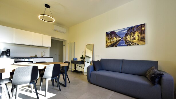 A modern living room with a sofa, dining table, and chairs. There is a kitchen area with cabinets and appliances. A painting of a cityscape is on the wall. - Studio Donatello Flat (Milano)