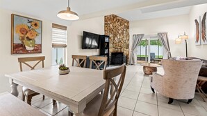 A dining area with a wooden table and chairs, a stone fireplace, a TV mounted on the wall, and a large window with curtains. -  Saltwater Pool I Firepits I Large Fenced Backyard I 4 Miles to Beach (Jacksonville)