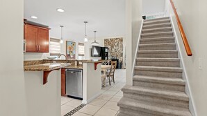 A modern kitchen with granite countertops, wooden cabinets, and a built-in dishwasher. A dining area with a table and chairs is visible in the background. A staircase with a wooden handrail leads to an upper floor. -  Saltwater Pool I Firepits I Large Fenced Backyard I 4 Miles to Beach (Jacksonville)