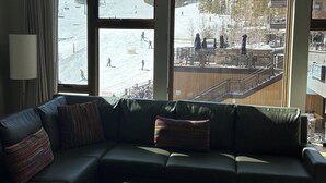 Een moderne woonkamer met een groot raam met uitzicht op een besneeuwde omgeving, een leren bank met kleurrijke kussens en een houten salontafel.