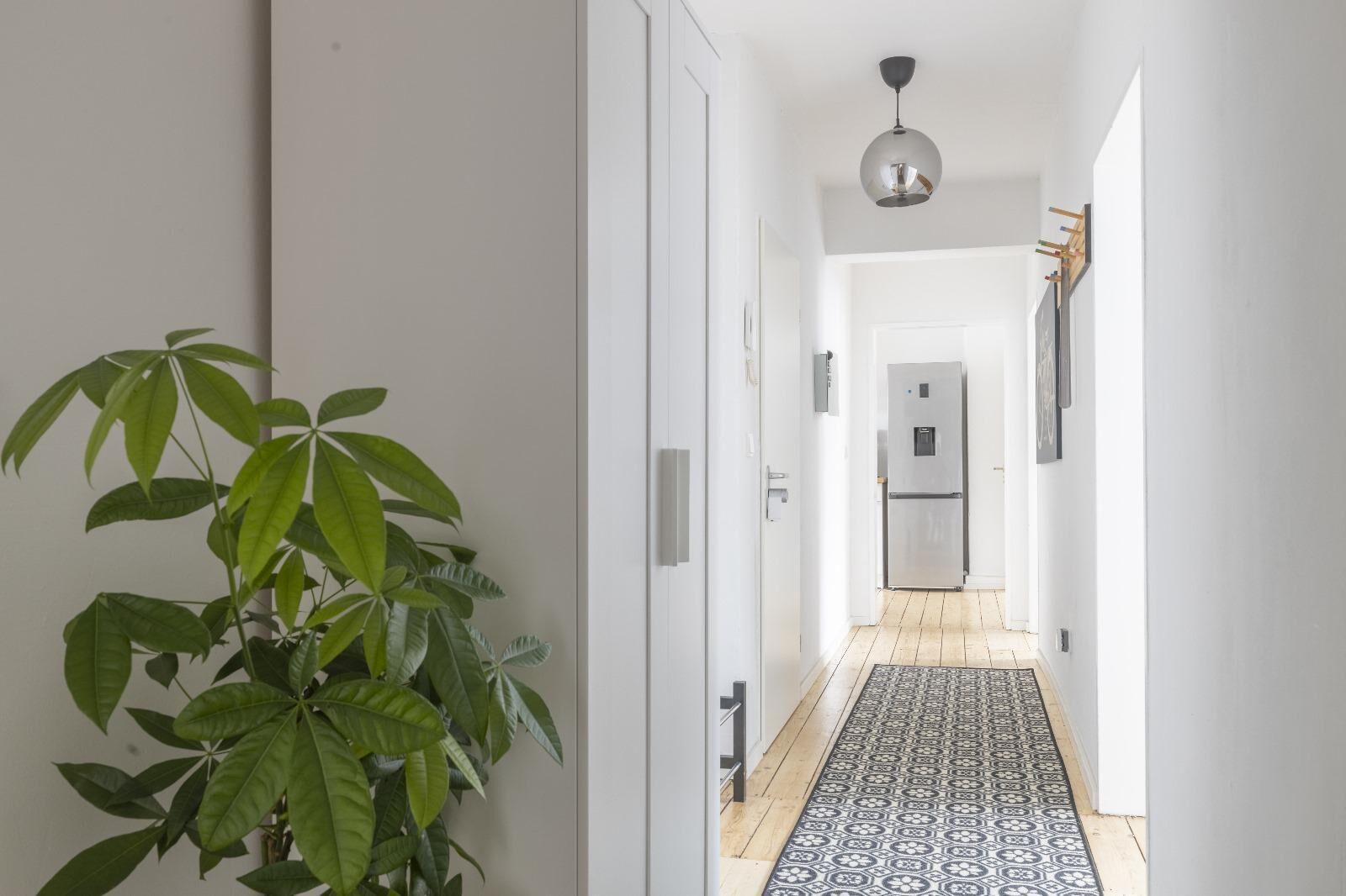 A hallway with a potted plant, a patterned rug, a refrigerator, and a pendant light.