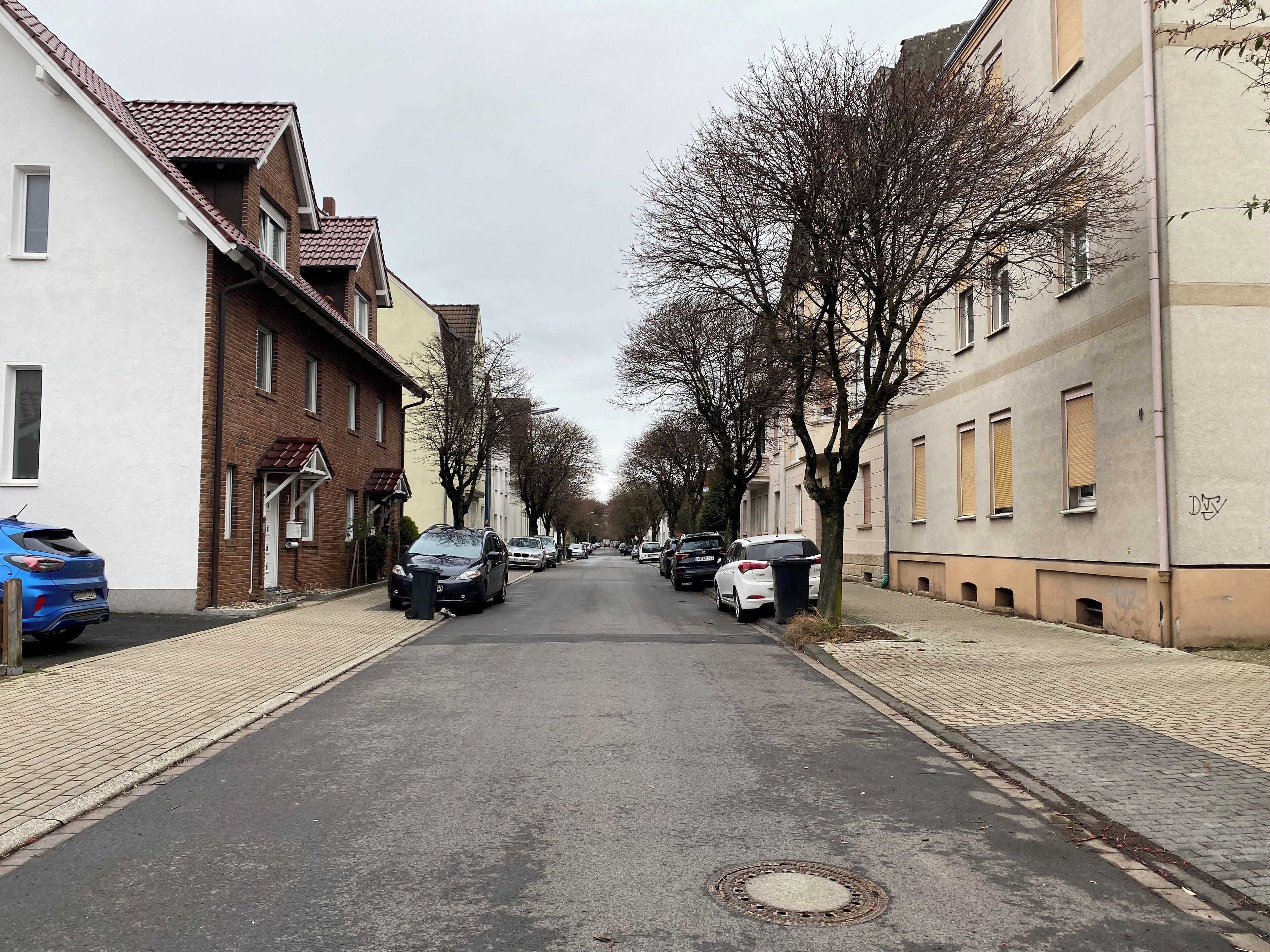 A narrow urban street lined with residential buildings, parked cars, and leafless trees.