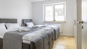 A modern bedroom with two beds, a grey headboard, white bedding, a window with blinds, and a radiator. - Gemütliches wohnen in idealer Lage, nahe Dortmund! (Holzwickede)