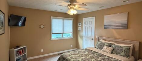 A bedroom with a bed, a desk, a chair, a ceiling fan, a window, and a television.