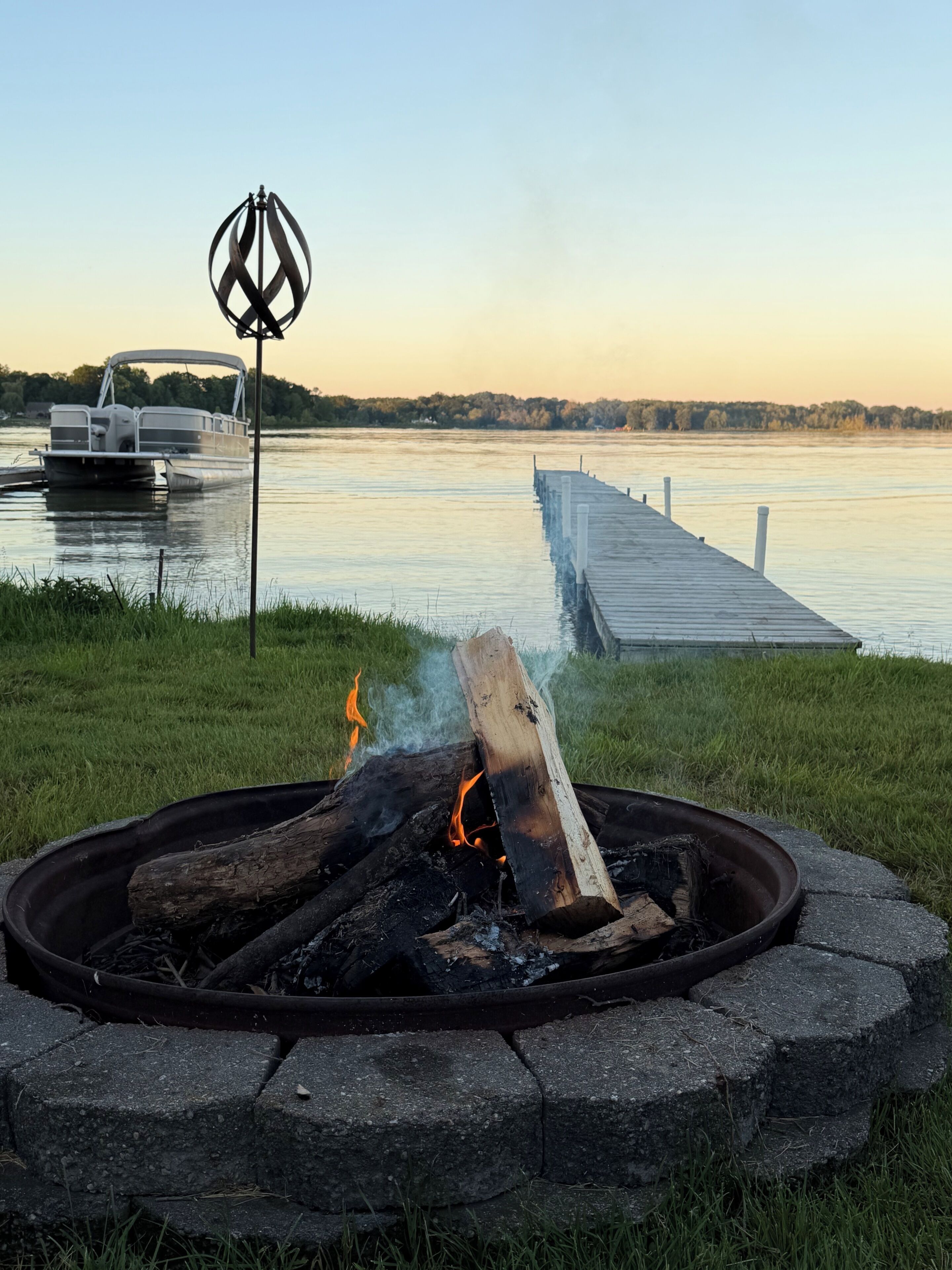 Ein Lagerfeuer mit Holzscheiten, ein Steg, der sich in einen ruhigen See erstreckt, und ein Boot im Hintergrund.
