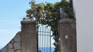 Une entrée fermée par un portail, avec un mur en pierre et un portail en métal, qui mène à un bâtiment à l’extérieur blanc et doté d’un toit recouvert de tuiles.