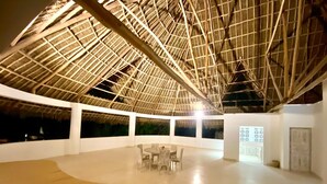 Un espacio interior con techo alto, estructura de techo de paja y una zona central de estar con mesas y sillas.