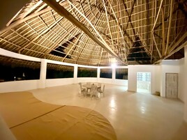 An indoor space with a high ceiling, a thatched roof structure, and a central seating area with tables and chairs.
