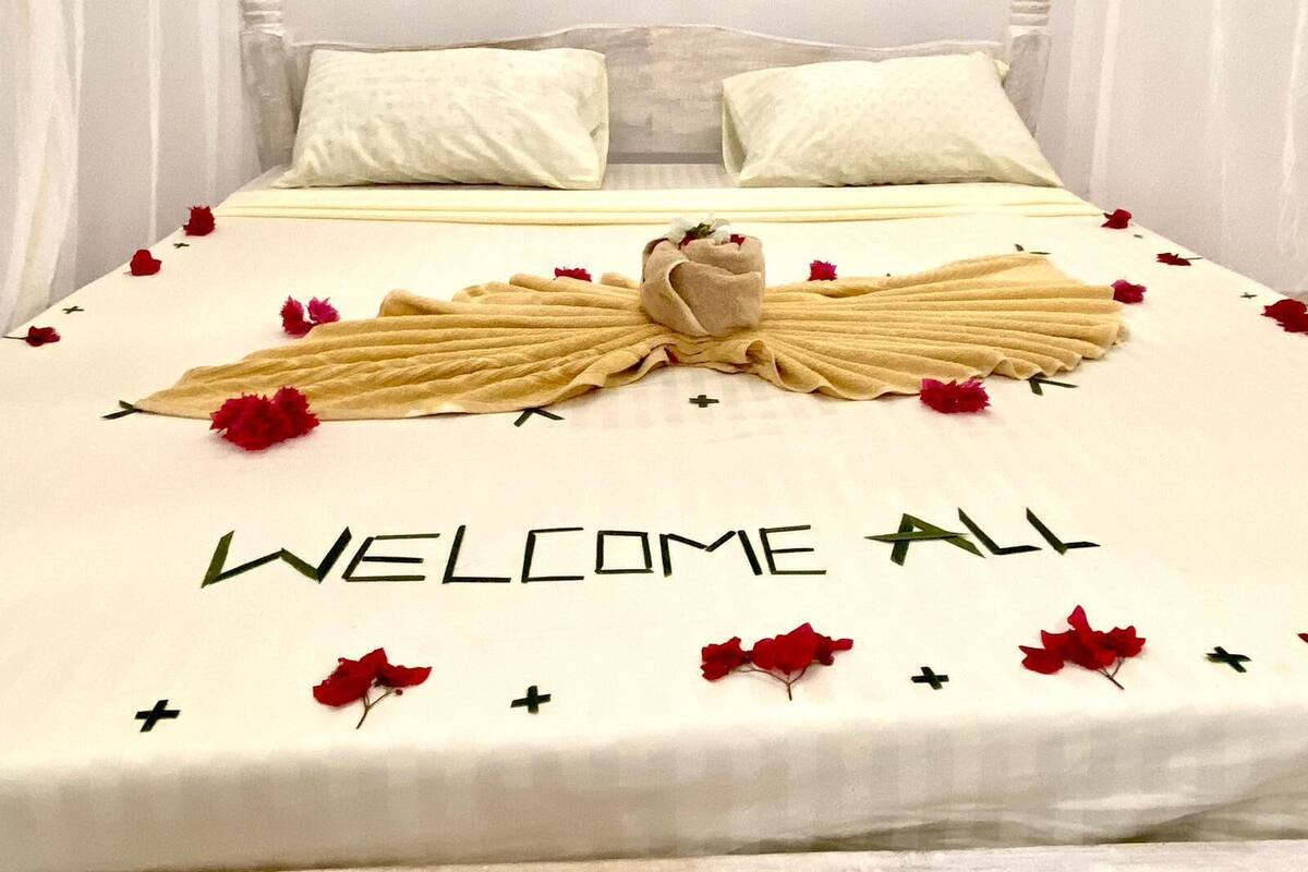 A bed with a towel arranged in the shape of a bird, with the words "WELCOME ALL" and red flowers scattered around.