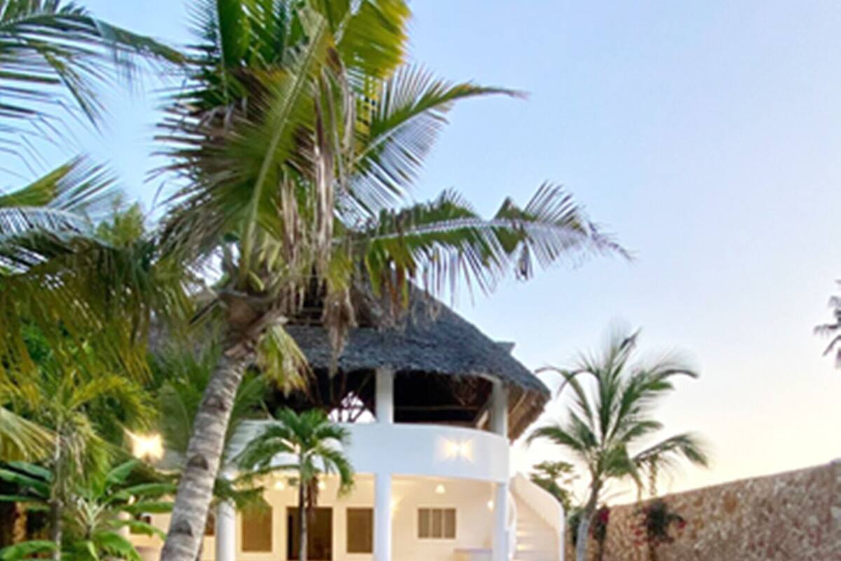 A modern resort with a pool, palm trees, and a thatched-roof structure.