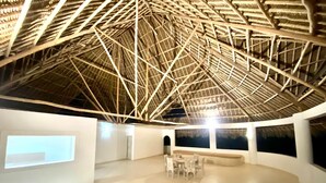 Amplia habitación con techo alto que presenta una estructura de cerchas de madera. Cuenta con un área de estar con mesas y sillas, y ventanas grandes a lo largo de las paredes.