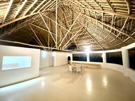 A spacious room with a high ceiling featuring a wooden truss structure. There is a seating area with tables and chairs, and large windows along the walls.