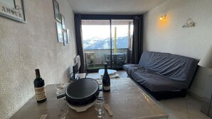 Quarto pequeno com sofá, mesa com grelha, garrafas de vinho e copos. Possui uma varanda com vista para as montanhas.