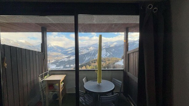 Uma varanda com mesa e cadeiras, guarda-corpo de madeira e vista para montanhas nevadas.