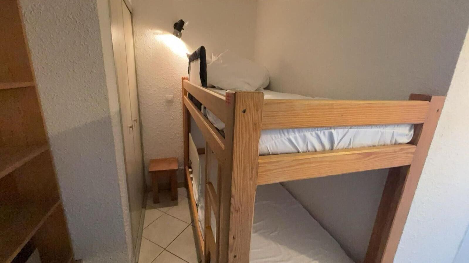 Een kleine kamer met een enkel bed, een houten stapelbed, een klein krukje en een wandlamp.