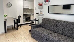 Una sala de estar moderna con un sofá gris estampado, un comedor con una mesa y sillas negras, una cocina con armarios blancos, un reloj y obras de arte en las paredes.