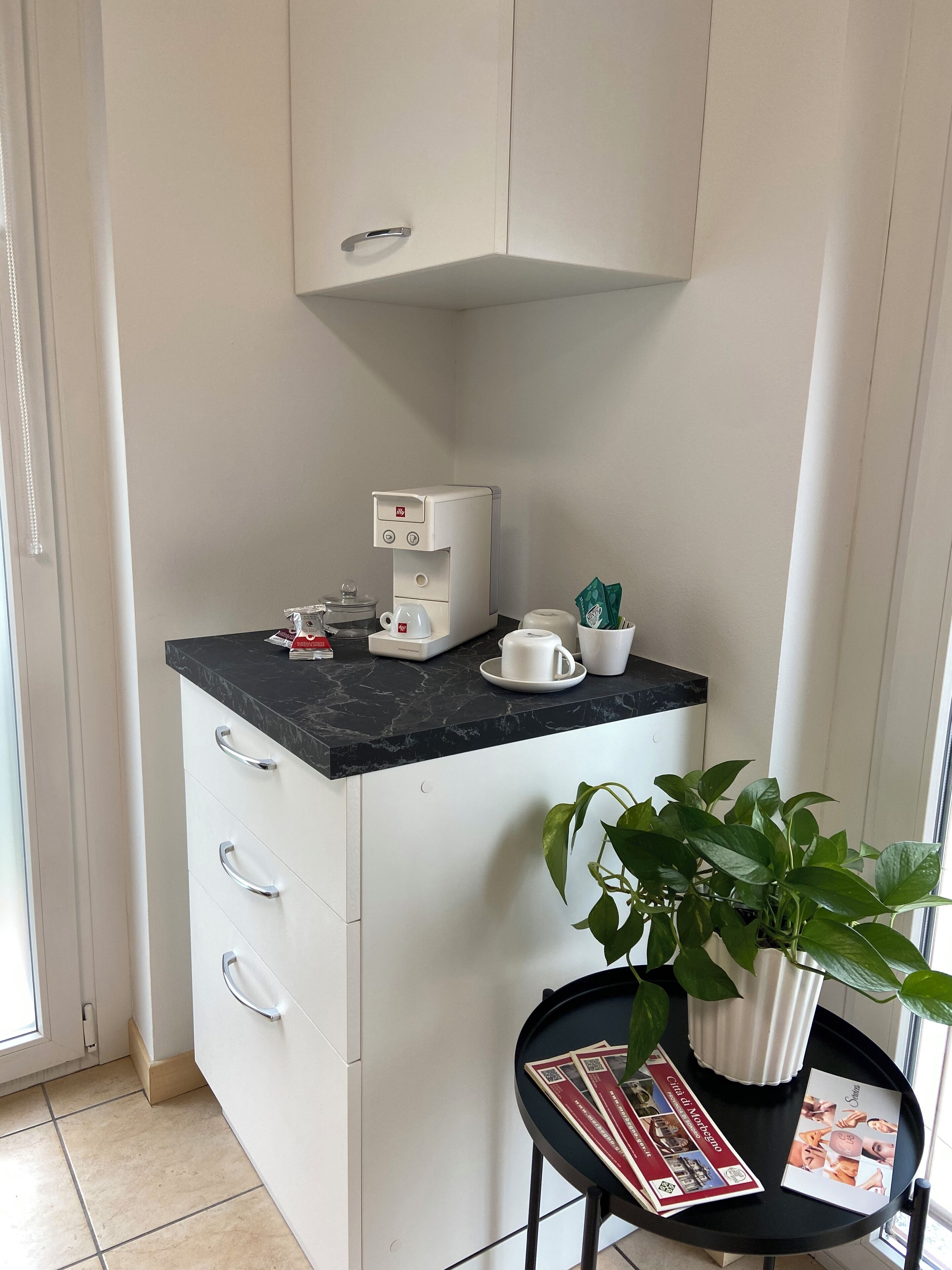 A compact kitchen with a coffee maker, a potted plant, and a magazine on a small table.