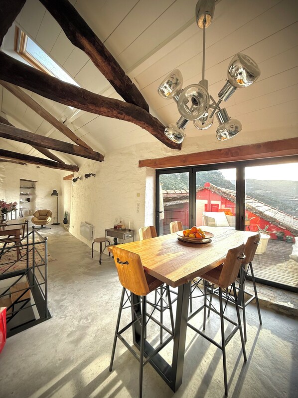 A spacious dining area with a large wooden table, modern chairs, and a view of a red-roofed building through large windows. - Ostal de la Croix Vieussan, Southern France (Vieussan)