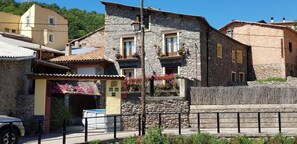 A stone building with a balcony, a small shop with a yellow awning, and a paved street with a metal fence.