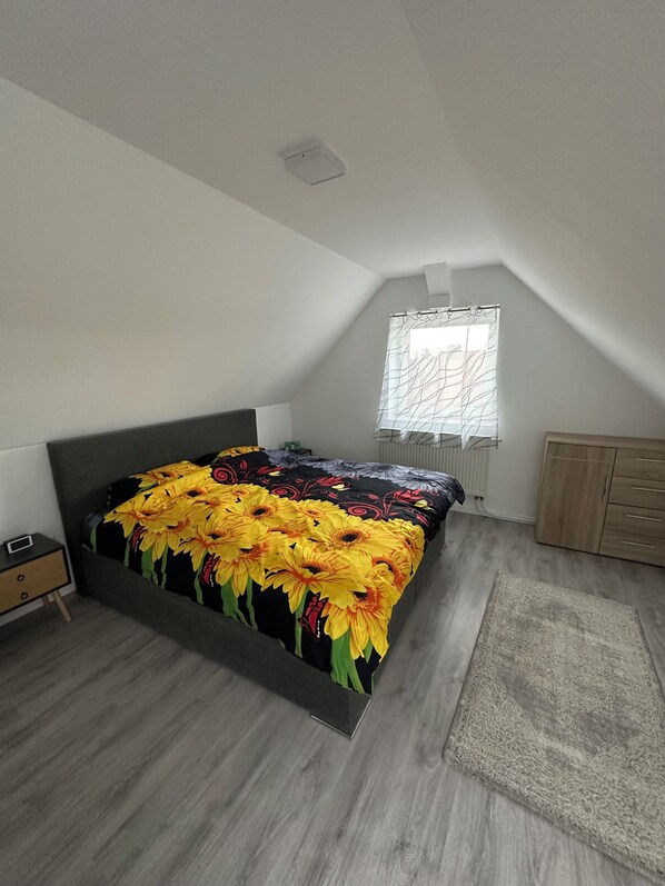 Une chambre à coucher comprenant un lit, une table de chevet, une commode, une fenêtre avec des rideaux et un radiateur.