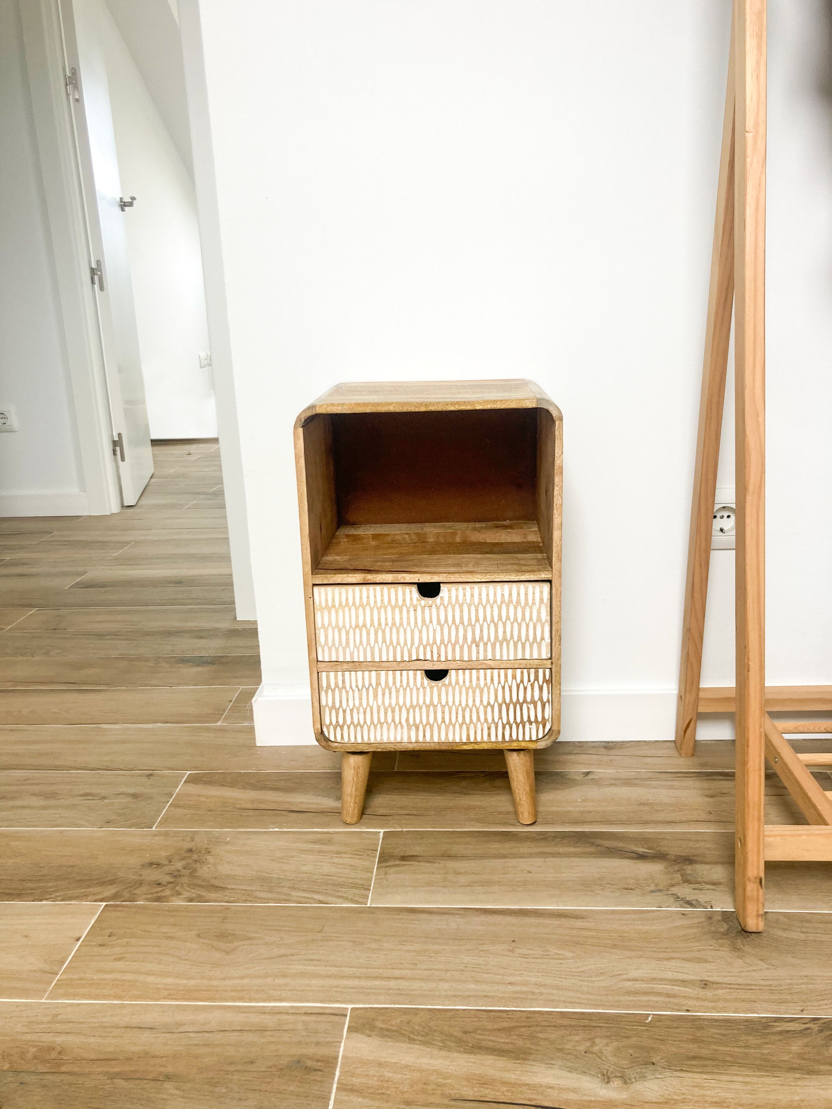A wooden bedside table with drawers against a white wall, wooden flooring, and a door in the background.