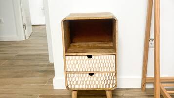 A wooden bedside table with drawers against a white wall, wooden flooring, and a door in the background.