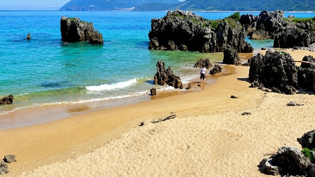 Ein Strand mit klarem türkisblauem Wasser, Felsformationen und Bergen im Hintergrund.