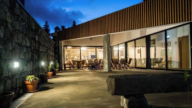 A modern building with a stone wall on one side, a wooden slatted roof, and a glass entrance leading to an outdoor seating area with tables and chairs.