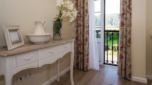 A room with a white console table, a vase with white flowers, a picture frame, and a view of a balcony with floral curtains.