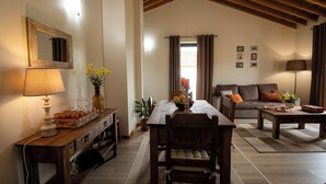 A cozy living room with a wooden dining table, a sofa, and a coffee table. There is a lamp, a vase with flowers, and a potted plant.
