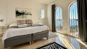 Une chambre à coucher avec un lit, une table de chevet, un tableau et une vue dégagée sur l’extérieur grâce à de grandes fenêtres.