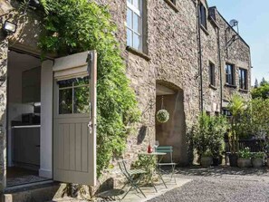 A stone building with a door open to a kitchen, a hanging wreath, and a table with chairs outside. - Cosy and characterful cottage in the heart of Kirkby Lonsdale (Kirkby Lonsdale)