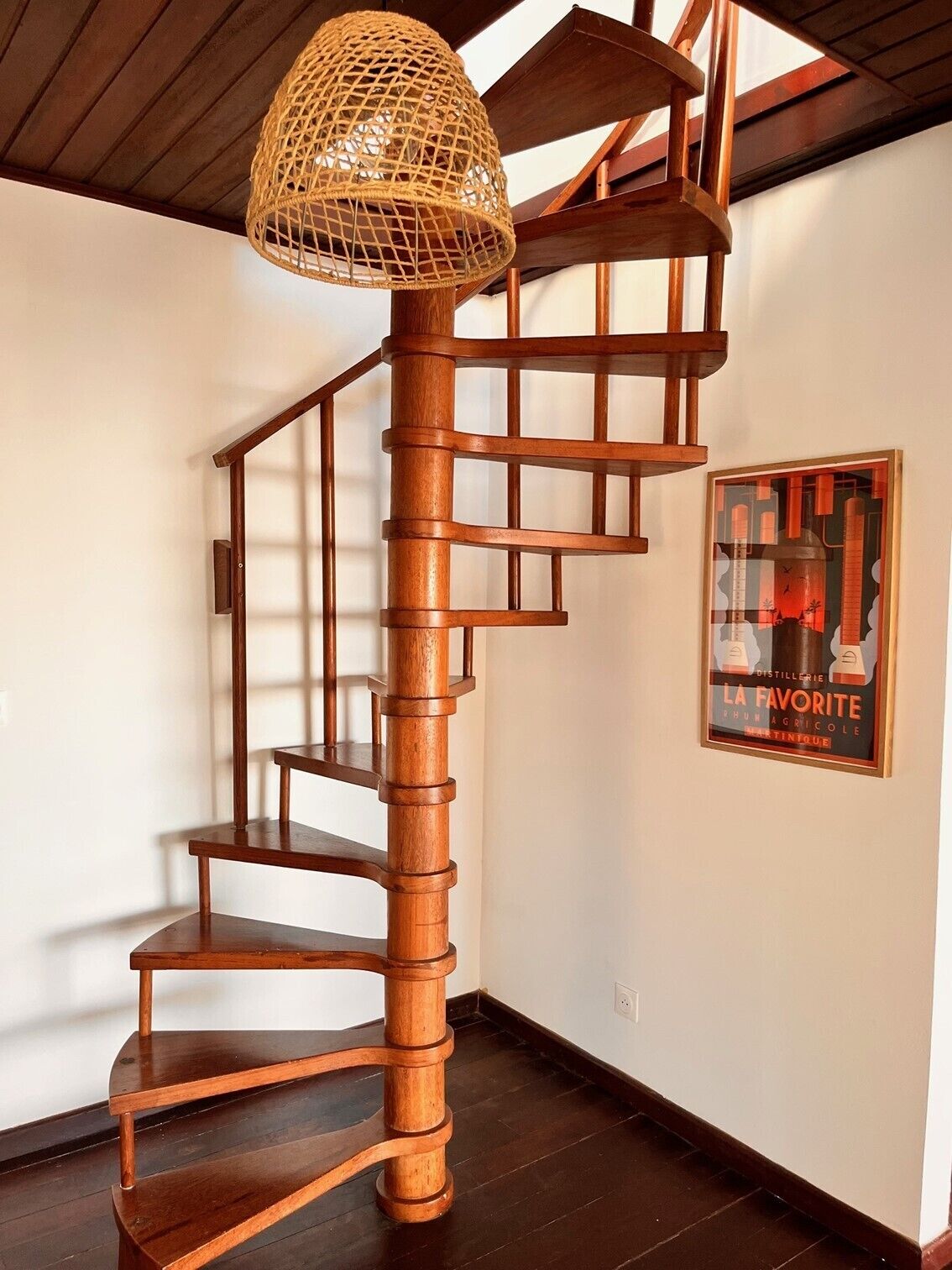 Un escalier en colimaçon en bois, surmonté d’un abat-jour tressé, une affiche encadrée accrochée au mur et un plancher en bois foncé.