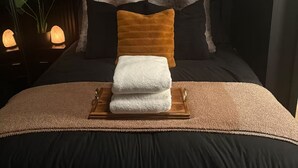 Een net opgemaakt bed met een houten dienblad waarop twee opgevouwen handdoeken liggen, een donker hoofdbord en een wandlamp.