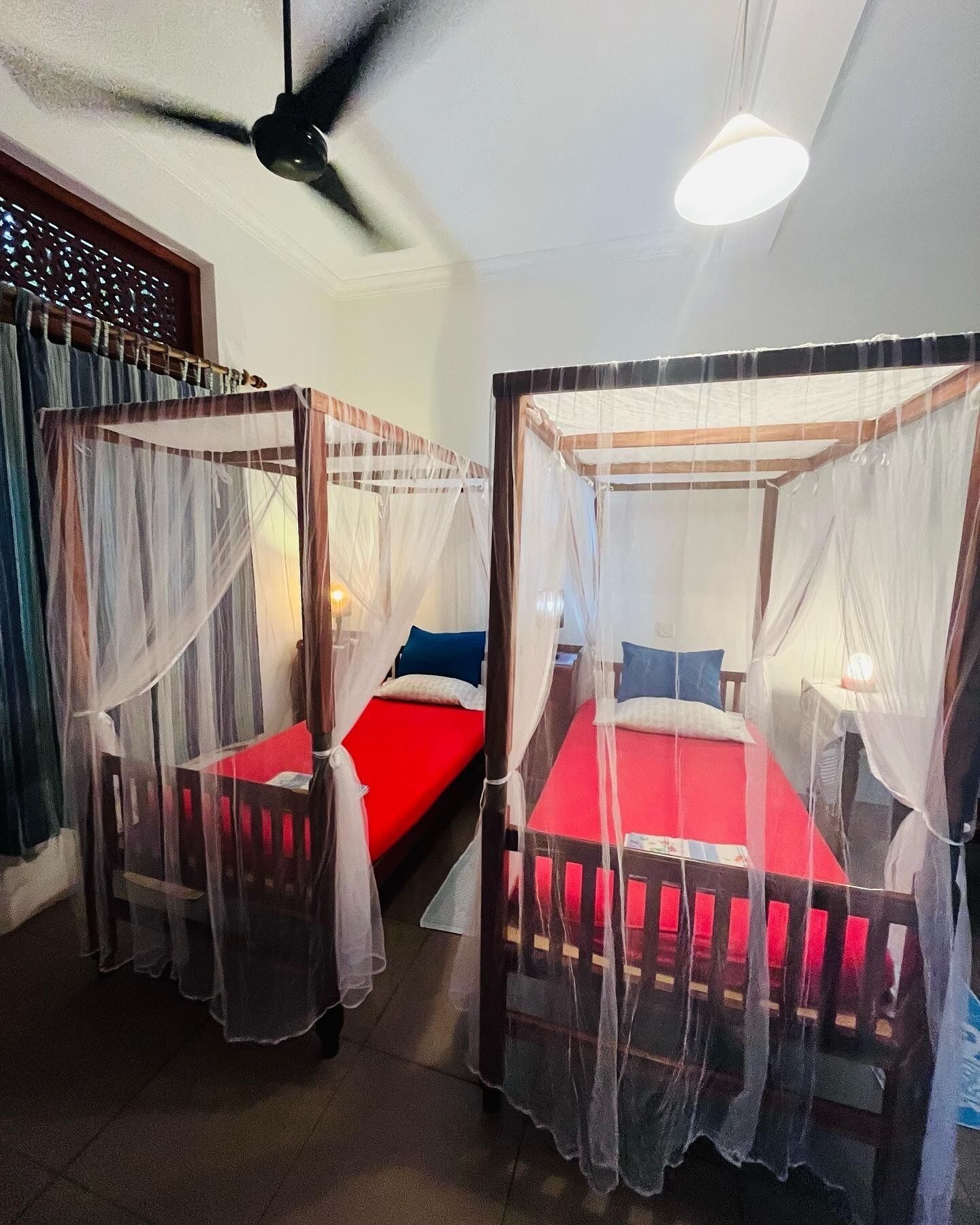 A room with two beds, each with a canopy. There is a ceiling fan and a wall-mounted light fixture.