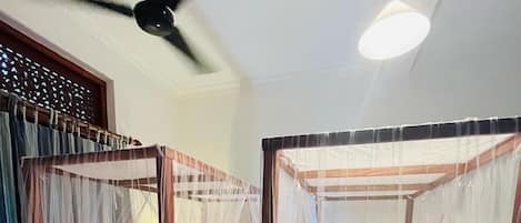 A room with two beds, each with a canopy. There is a ceiling fan and a wall-mounted light fixture.