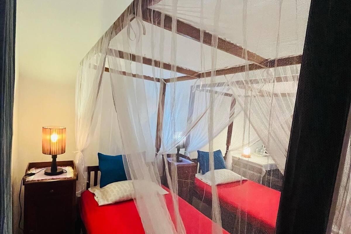 A room with a bed under a mosquito net, a nightstand with a lamp, and a red bedspread.