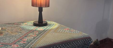 Una habitación con una mesa cubierta por un mantel estampado, una lámpara con pantalla cilíndrica y una silla de madera con respaldo curvo.