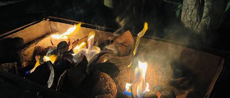 A fire pit with burning logs surrounded by rocks, set in a dark outdoor area with some vegetation in the background.