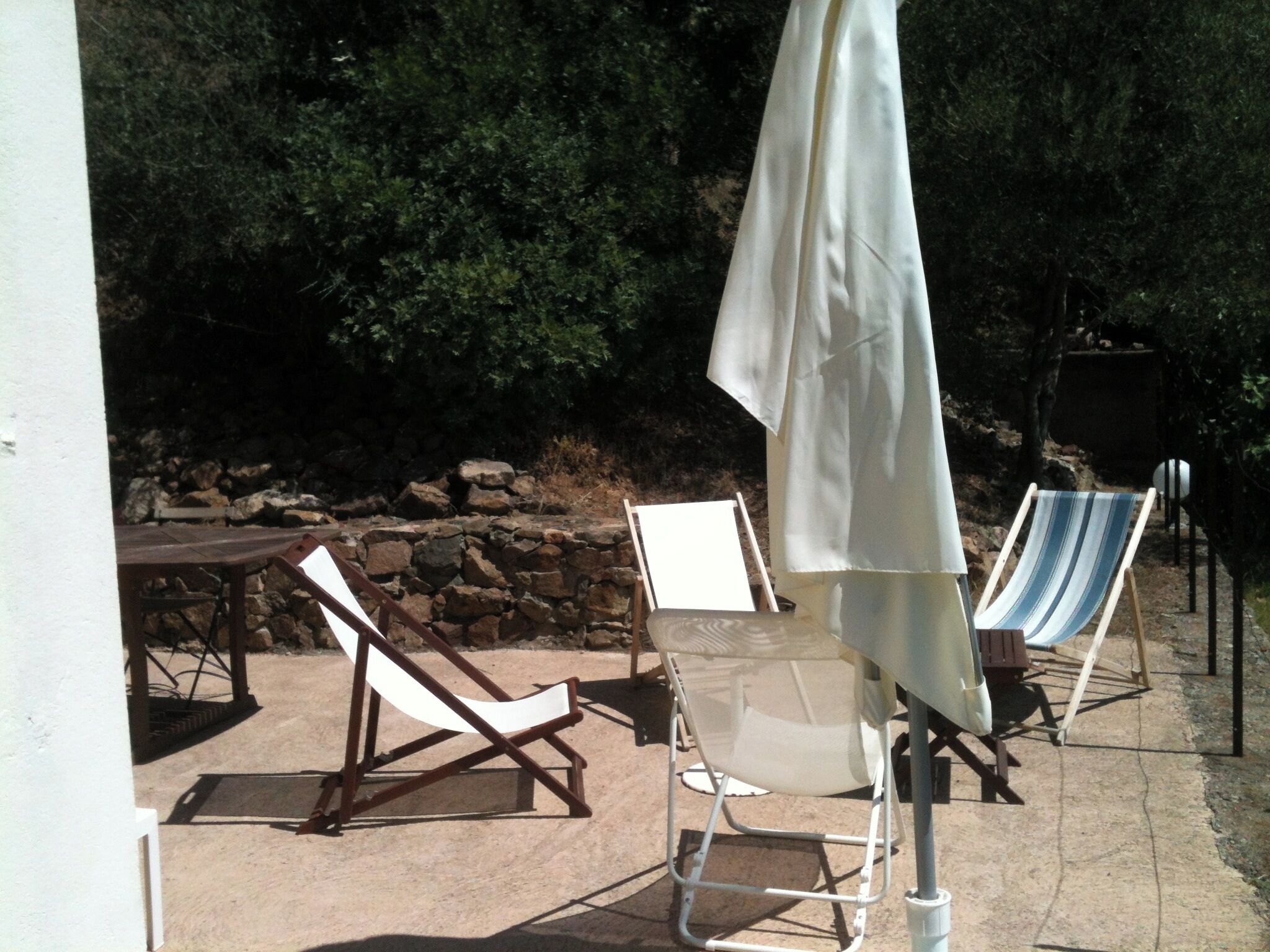 Un área de estar al aire libre con dos sillones, una mesa, un paraguas blanco y un muro de piedra al fondo.