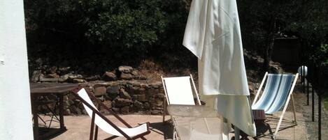 Un área de estar al aire libre con dos sillones, una mesa, un paraguas blanco y un muro de piedra al fondo.