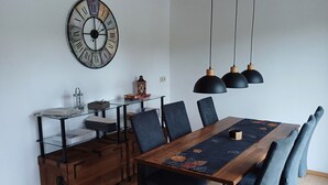 Comedor con mesa de madera, sillas negras, un reloj grande en la pared, luces colgantes y una mesita auxiliar.