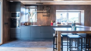 A modern kitchen with dark cabinetry, a central island, bar stools, and a hanging light fixture. - Apartment with private pool in Annecy (Annecy)