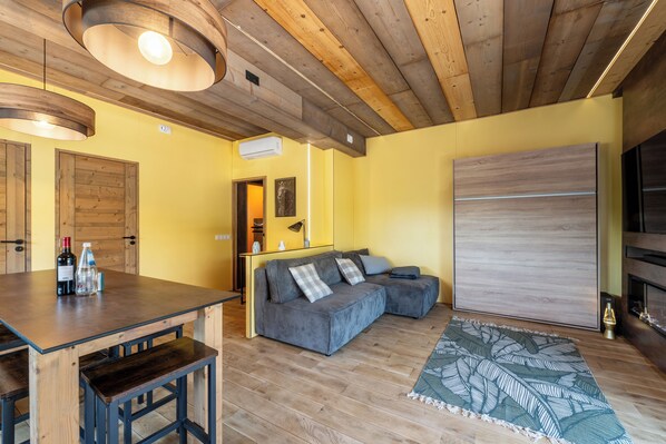 A cozy living area with a wooden ceiling, a grey sofa with cushions, a wooden dining table with bottles, and a patterned rug.